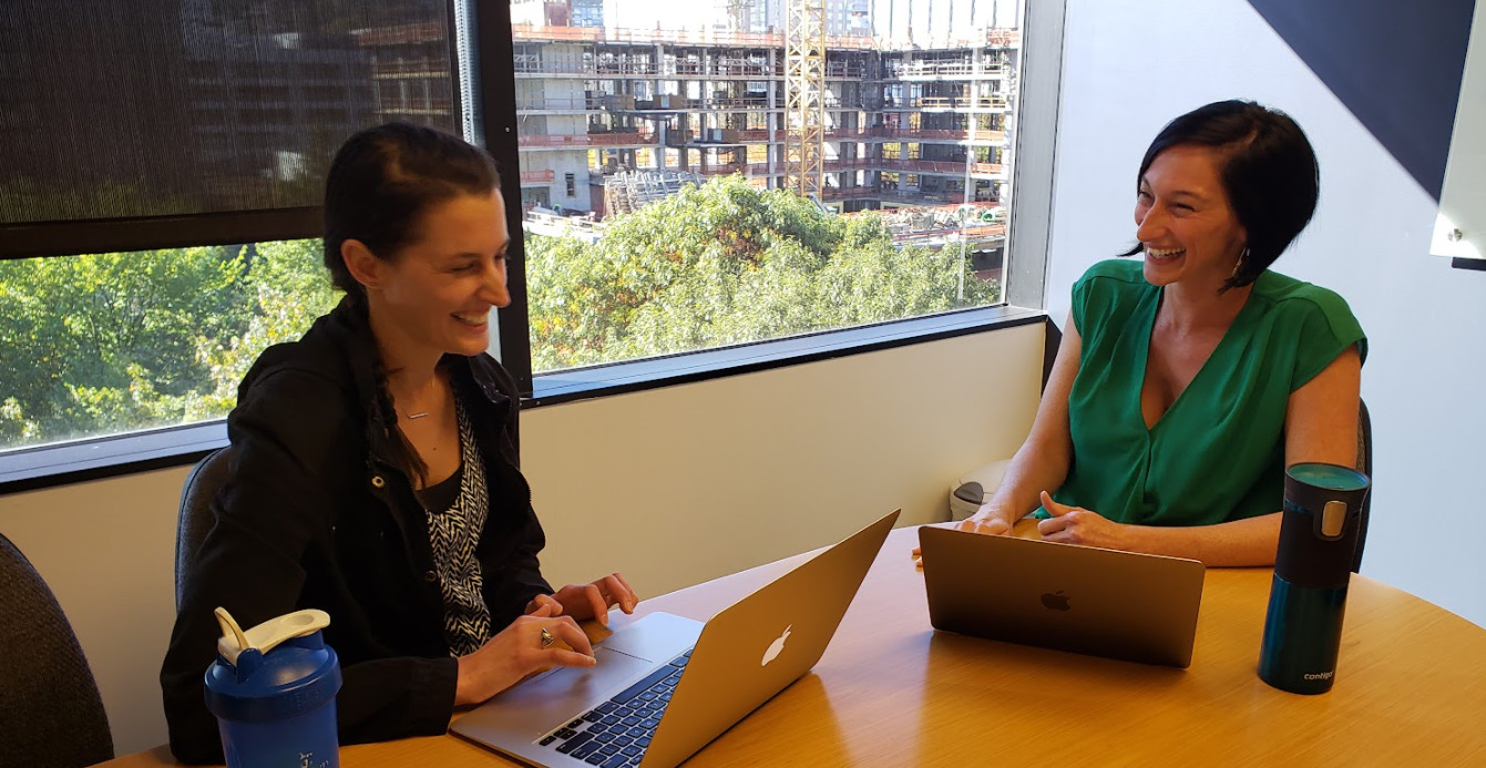 a female manager wearing a green shirt smiling while chatting with female employee wearing a black sweater also smiling during coaching meeting